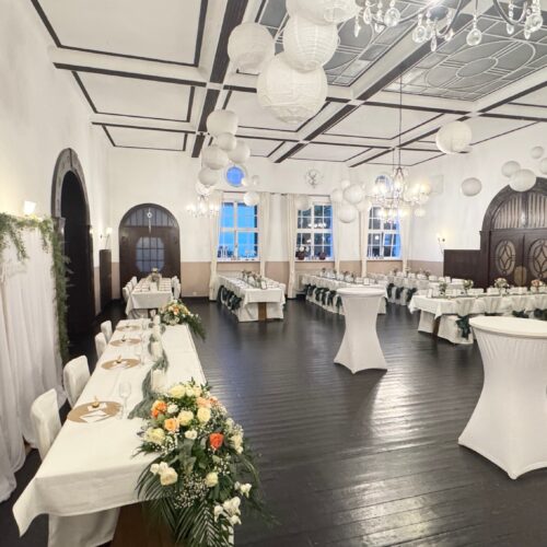 Großer Festsaal mit weißen Tischdecken, Blumenarrangements und runden Lampen.