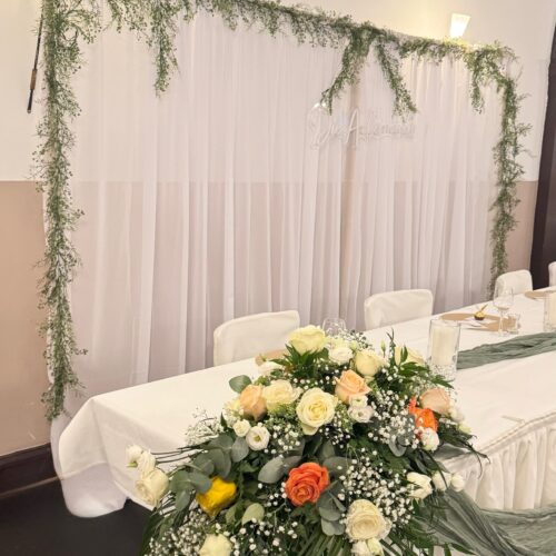 Weiß gedeckter Hochzeitstisch mit Blumengesteck, Vorhänge im Hintergrund.